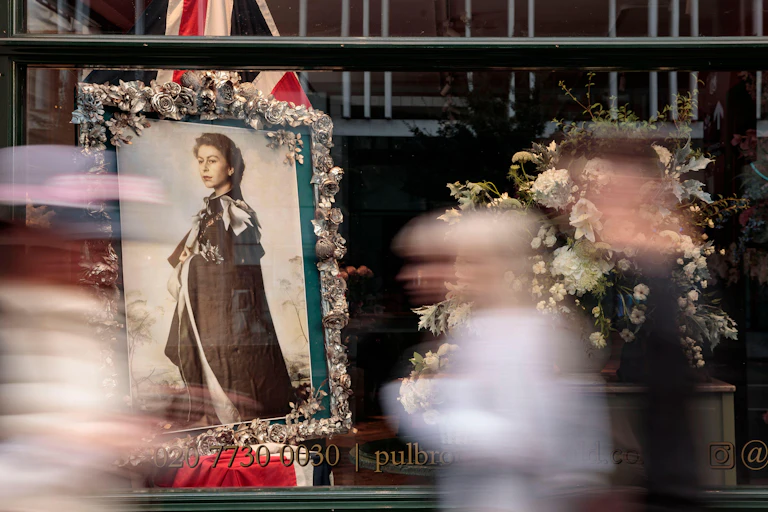 A photograph of a photograph of the Queen in a shop window with people passing fast, their silhouettes all blurred.