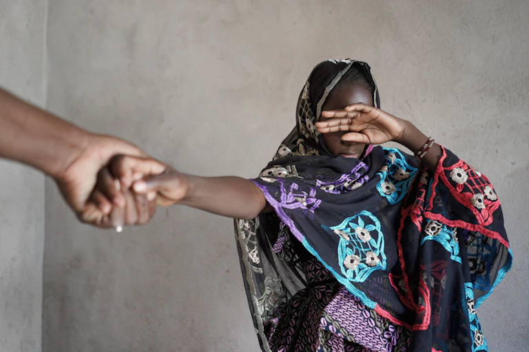 A young woman of African ethnicity wearing a black dress decorated with blue, red and purple patterns covers her eyes with one hand while an adult hand is seen pulling her by the hand