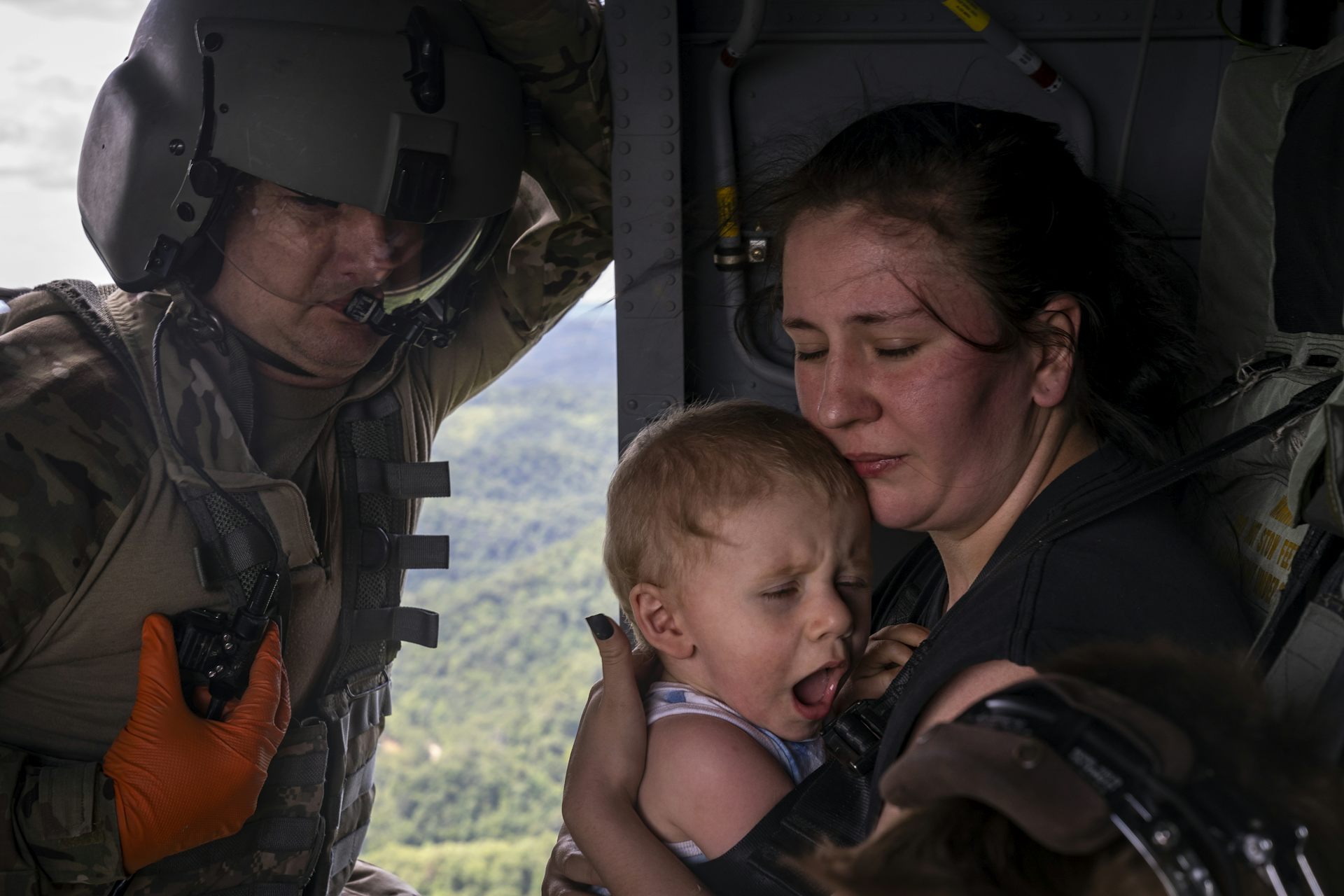 A woman with her eyes closed holds a screaming 1-year-old boy in a National Guard helicopter, with a guard member standing in the open helicopter door.
