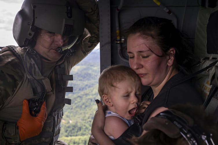 A woman with her eyes closed holds a screaming 1-year-old boy in a National Guard helicopter, with a guardsman standing in the open helicopter door.