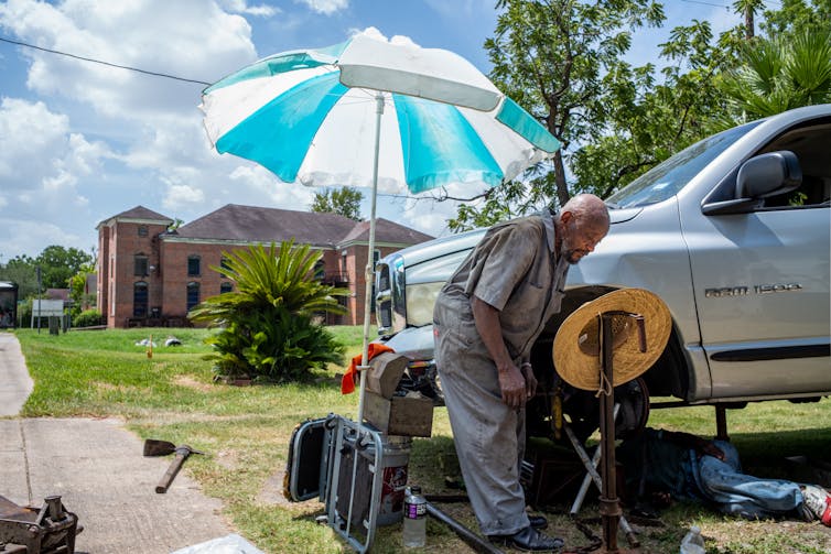 A man is working on a car, with an elderly mechanic in overalls standing next to him in the shade of a large parasol.