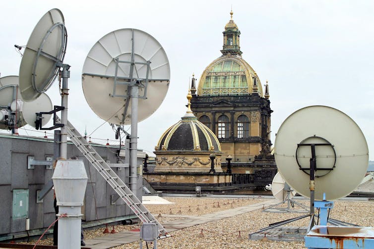 Satellite dishes adorn a roof, with church spires in the background