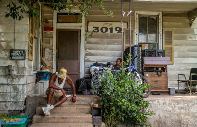 A 62-year-old man sits on the steps of a house in 102 degree heat during a July heat wave in Texas. Another person sits on the porch.