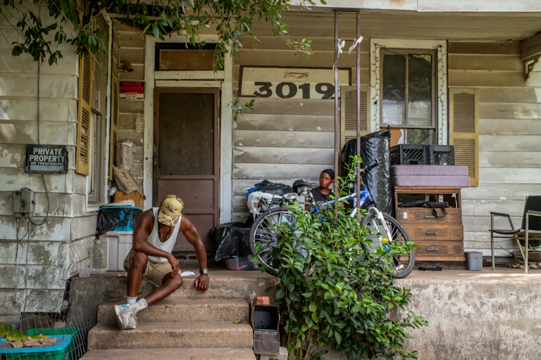 A 62-year-old man sits on the steps of a house in 102 degree heat during a July heat wave in Texas. Another person sits on the porch.