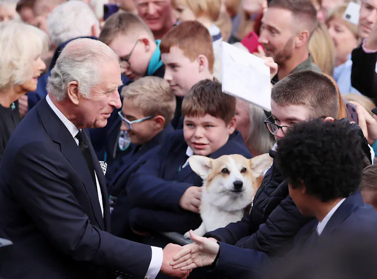 King Charles shaking hands with a young man as a corgi looks on.