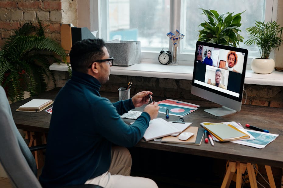 An instructor sits on a chair and students are seen on a videoconferencing screen.