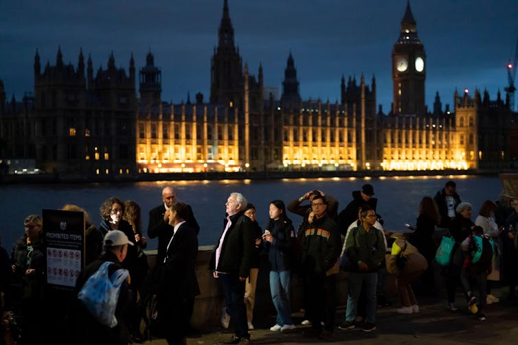 A queue of people along London's south bank at night, with the Palace of Westminster illuminated in the background across the river.