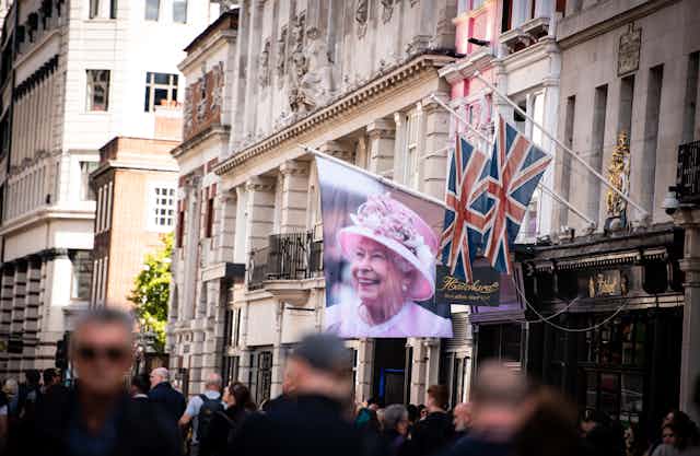 A flag with a photo of the late Queen hangs on the side of an ornate building while hundreds of people walk past