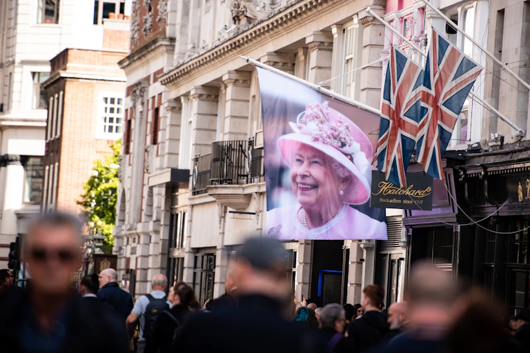 A flag with a photo of the late Queen hangs on the side of an ornate building while hundreds of people walk past