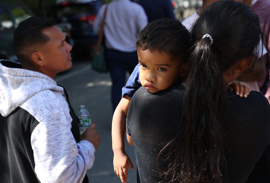 A small boy is seen resting his head on his mother's shoulder, while she stands next to several Latino people.