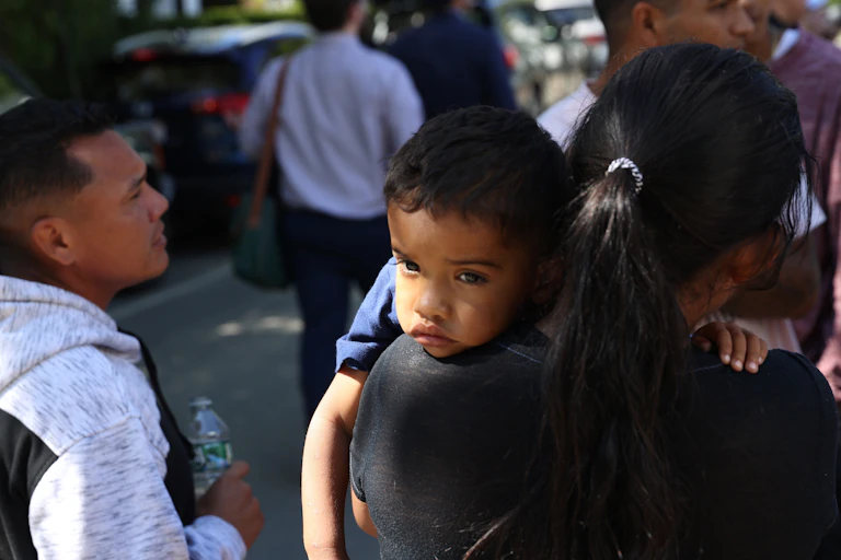 A small boy is seen resting his head on his mother's shoulder, while she stands next to several Latino people.