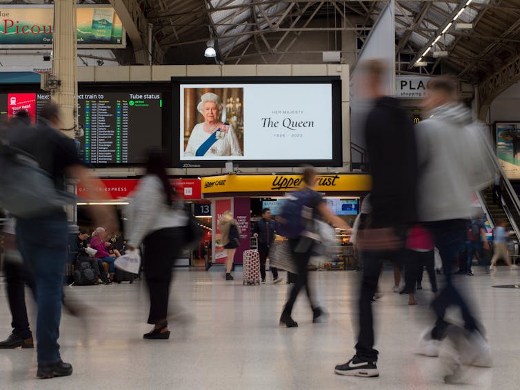 A memorial image of the Queen is displayed on a screen in a train station, while commuters file past