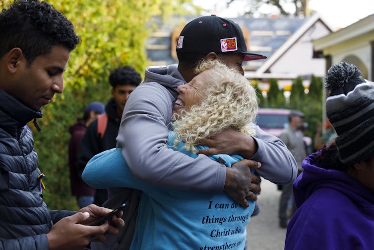 A blond curly-haired woman hugs a Latino young man wearing a baseball cap.