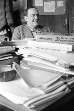 A man in a suit sits behind a desk covered in books and papers.