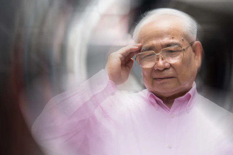 A man with gray hair wearing glasses with his hand to his forehead, with the foreground blurred