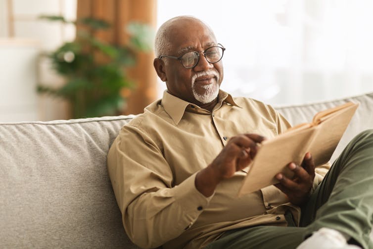 Bărbat în vârstă cu barbă care citește o carte așezată pe o canapea