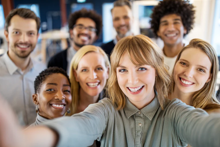 Eight smiling men and women, looking into a camera, take a group selfie.