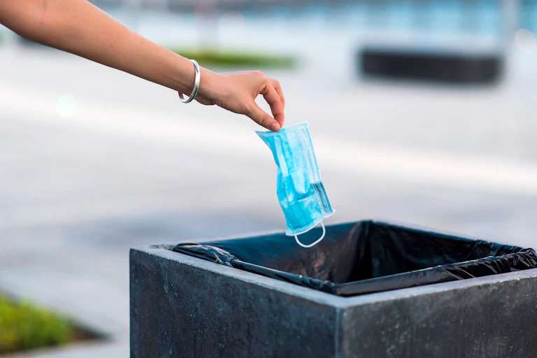 A hand dropping a surgical mask in a trash can.
