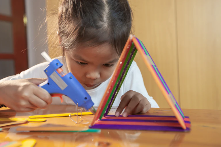 A child glues colourful paddle-pop sticks into a triangle with a glue gun.