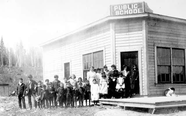 Black and white photo of children standing in front of a wooden building with sign that reads 'public school'