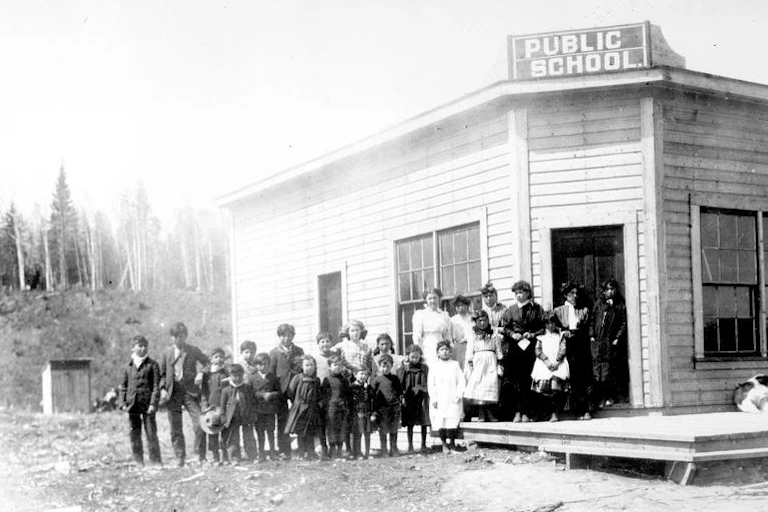 Black and white photo of children standing in front of a wooden building with sign that reads 'public school'
