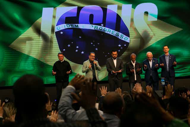 Six men pray on a stage in front of an image of the Brazilian flag.