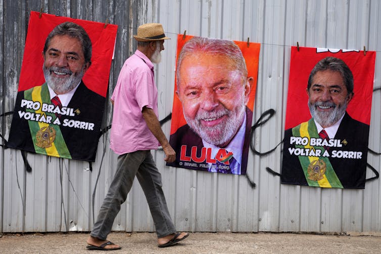 A man in a pink shirt and straw hat walks by three large campaign posters on a wall.