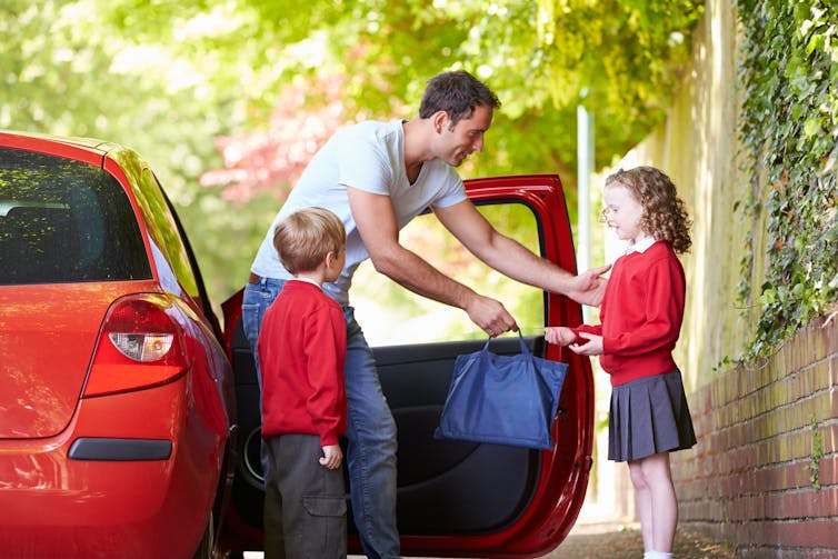 A man standing in front of a red car while dropping off two children at school.