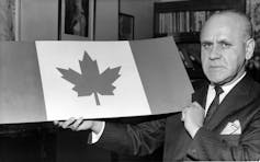 A black and white photo shows a bald man in a suit holding up the design of the new Canadian flag.