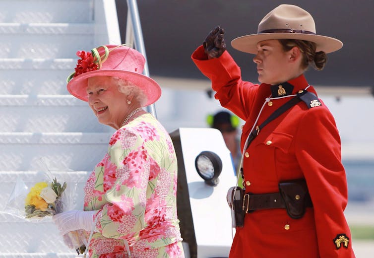 An elderly woman in a pink hat and floral dress, holding flowers, smiles as she boards a plane and is saluted by an RCMP officer.