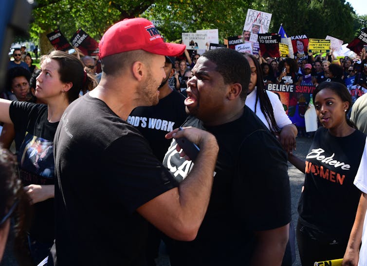 Um homem branco de chapéu vermelho parece estar discutindo com um jovem negro em uma cena lotada que parece um protesto.