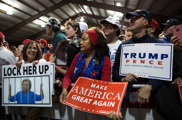 Women and men stand together with protest signs that say 'Make America Great Again' and 'Lock her up' at a Trump rally.