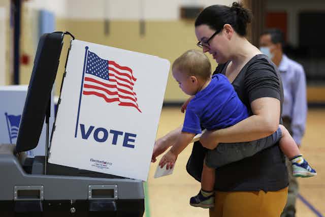 A white woman with dark hair holds a small child as they appear to vote, standing next to a white box that has an American flag on it and says 'vote.''