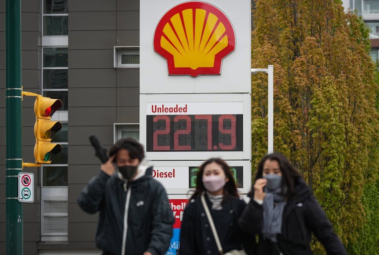 People walk in front of a Shell gas station sign displaying a gas price of 227.9