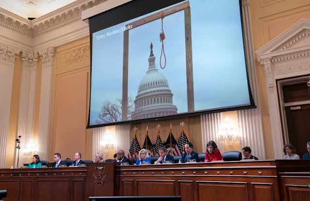 A large screen hangs above a panel, showing a photo of a gallows in front of the U.S. Capitol.
