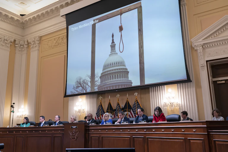 A large screen hangs above a panel, showing a photo of a gallows in front of the U.S. Capitol.