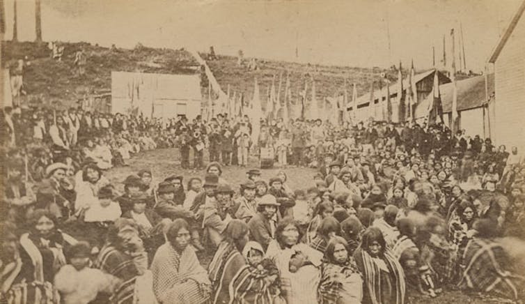An old photo shows a large group of people sitting outside in a large circle.