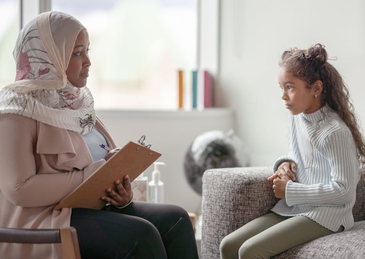A woman seated in an armchair with a clipboard and pen wearing a headscarf looks at a young girl with space buns who is looking back at her, seemingly pausing in conversation.