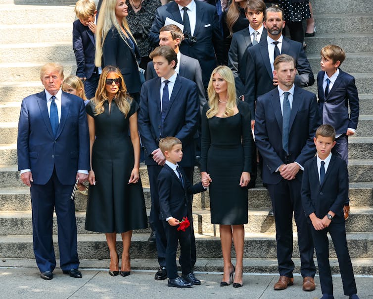 A group of people dressed in formal, dark clothing stand in front of white steps