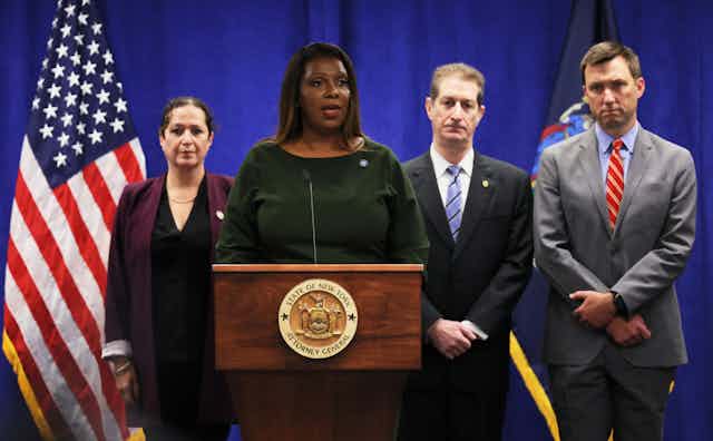 A black woman in a formal dress stands at a wooden podium, with three people and an American flag behind her.