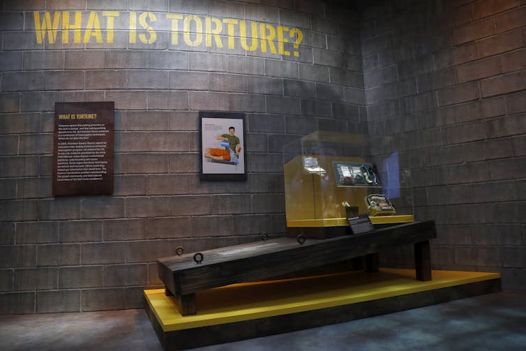 A museum display shows a wooden board the size of a person below the words 'What is torture?'