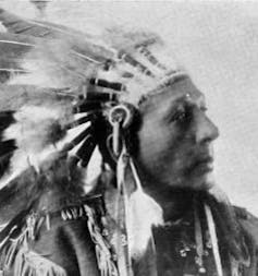 A black and white photo of a man wearing an Indigenous headdress of feathers.