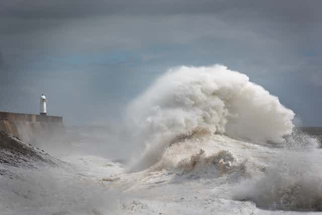 A giant wave dwarfs a lighthouse on a pier, against a grey sky.