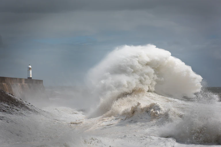 A giant wave dwarfs a lighthouse on a pier, against a grey sky.