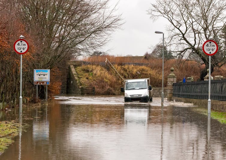 A van drives through a flooded street in a countryside setting.