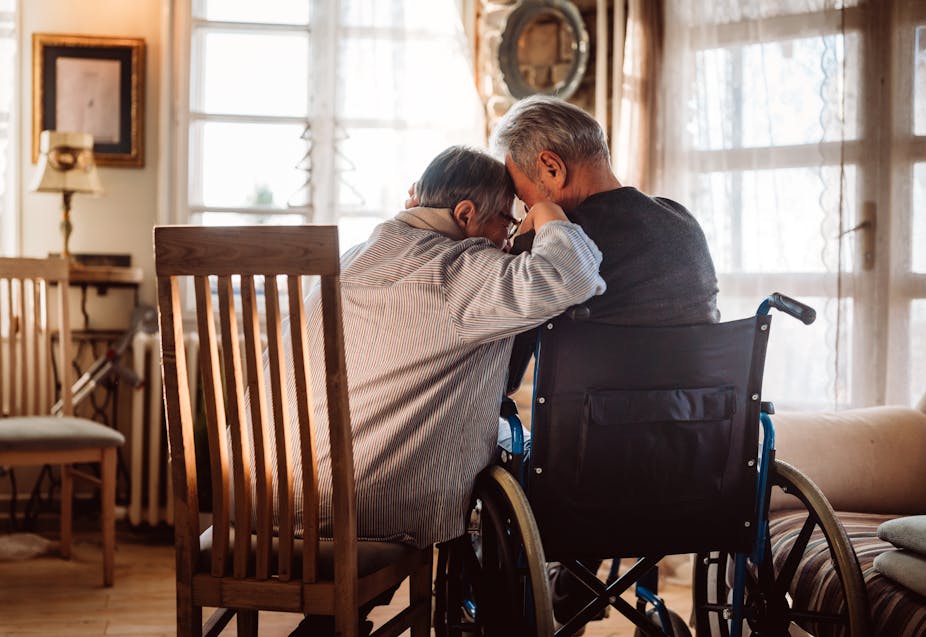 Photo from behind of an older couple consoling each other, the wife is leaning her head on the husband's shoulder, he is in a wheelchair and she is sitting in a chair