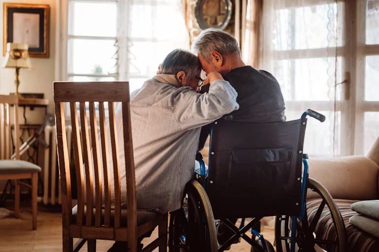 Photo from behind of an older couple consoling each other, the wife is leaning her head on the husband's shoulder, he is in a wheelchair and she is sitting in a chair
