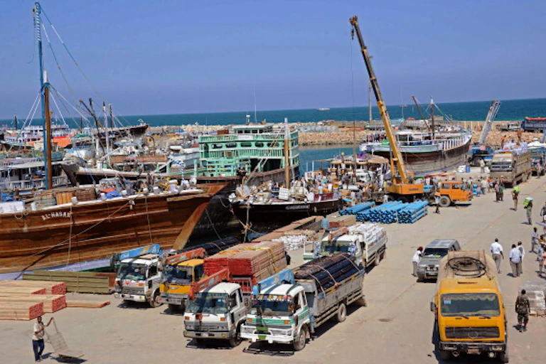 Five trucks loaded with long planks of wood and pipes waiting alongside ships docked at a port.