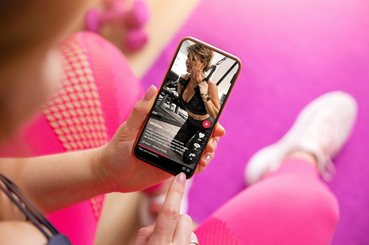 A woman in pink athletic gear holds a smartphone showing a TikTok video of another woman in black exercising