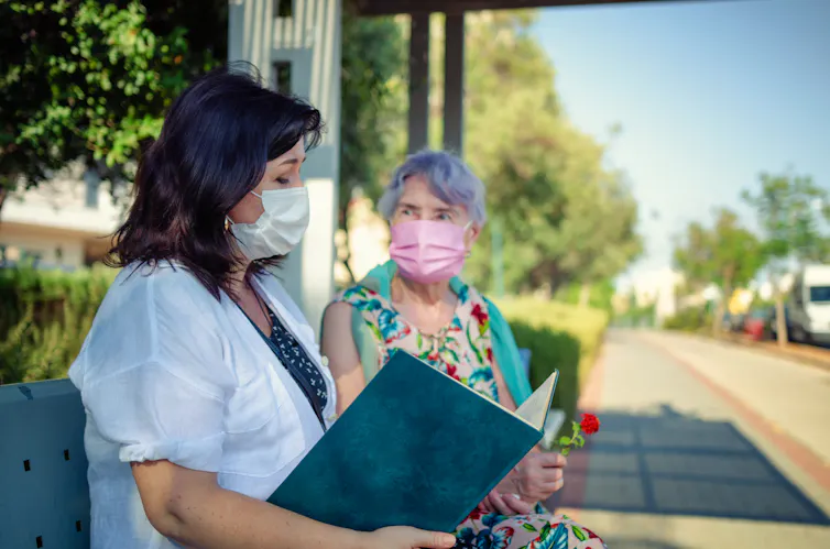 A middle age caregiver sitting and reading on a bench beside an elderly woman, presumably her client. The caregiver's face is in focus. The elderly woman is slightly blurred.
